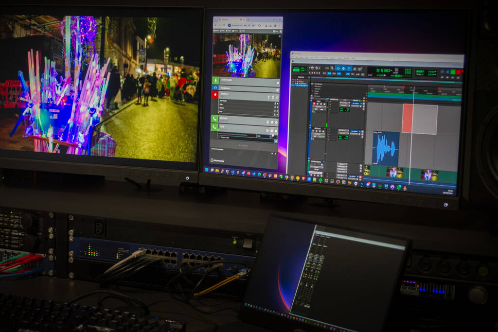 A dark production studio showing a television monitor with footage of a carnival alongside the ProTools audio editor and Cleanfeed