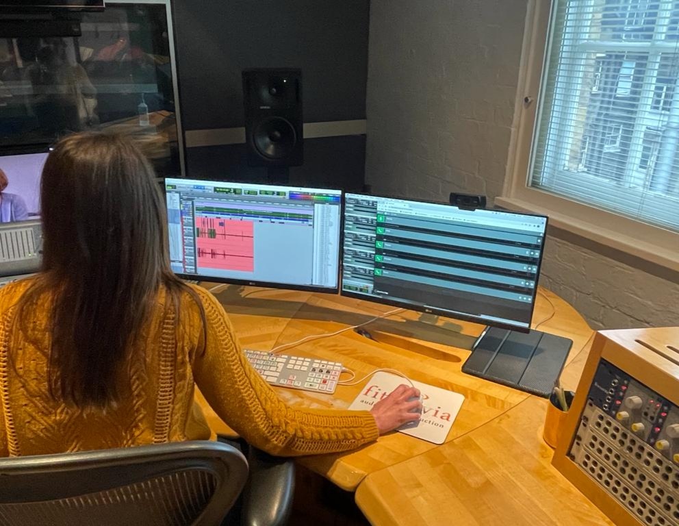 A woman in an orange sweater sits in front of screens running ProTools and Cleanfeed. Generlec speakers and a Fitzrovia Post mouse pad are visible